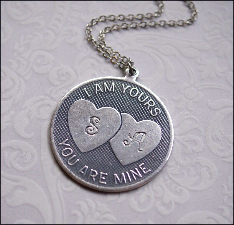 May include: A silver-toned pendant necklace with a circular design. The pendant features two heart shapes with the letters "S" and "A" inside. The words "I AM YOURS" and "YOU ARE MINE" are inscribed around the hearts. The necklace has a delicate chain.