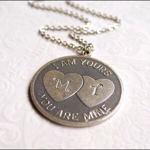 May include: Silver-toned pendant necklace with a round charm. The charm features two heart shapes with the letters "M" and "T" inside. The words "I AM YOURS" and "YOU ARE MINE" are engraved around the edge of the charm.