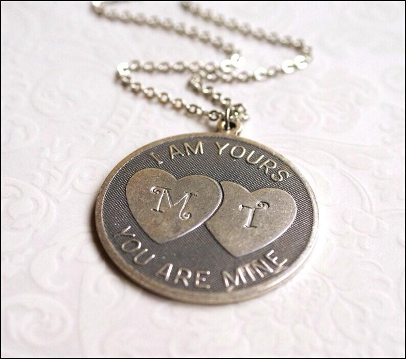 May include: Silver-toned pendant necklace with a round charm. The charm features two heart shapes with the letters "M" and "T" inside. The words "I AM YOURS" and "YOU ARE MINE" are engraved around the edge of the charm.