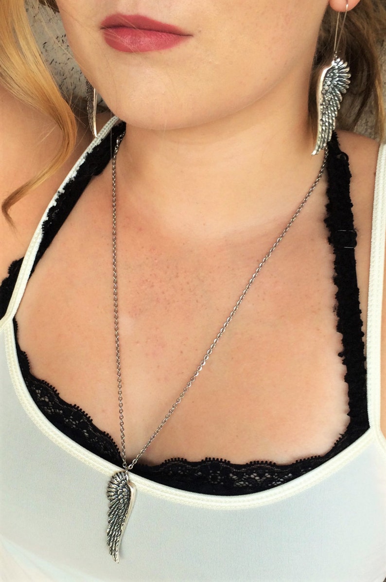 May include: A silver necklace with a wing pendant. The necklace is worn by a person with a white tank top and black lace trim.