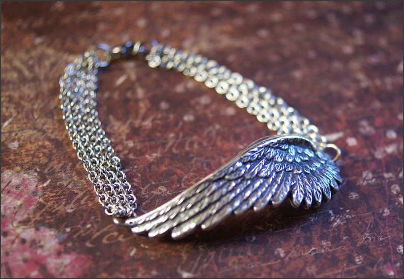 Puede incluir: Un brazalete de plata con un dije de ala detallada y m&uacute;ltiples eslabones de cadena. El brazalete est&aacute; sobre una superficie marr&oacute;n texturizada.