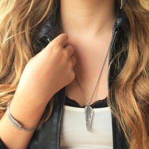 Puede incluir: Un collar de plata con un colgante de ala. El collar lo lleva una persona que lleva una chaqueta de cuero negra y un top blanco sin mangas.