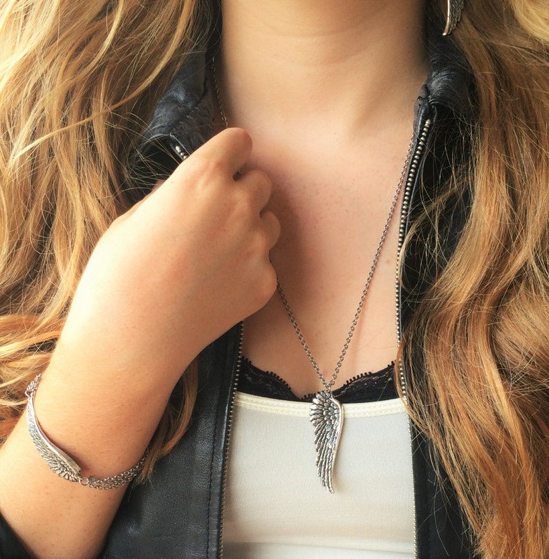 Puede incluir: Un collar de plata con un colgante de ala. El collar lo lleva una persona que lleva una chaqueta de cuero negra y un top blanco sin mangas.