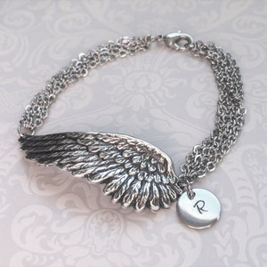 Puede incluir: Pulsera de plata con un dije de ala de &aacute;ngel detallado y un dije redondo con la letra "R" grabada.