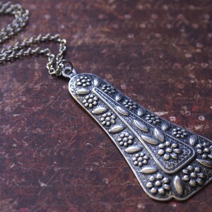 Puede incluir: Un collar con un colgante de plata con un dise&ntilde;o floral detallado. El colgante tiene forma de l&aacute;grima y presenta intrincados tallados de hojas y flores.