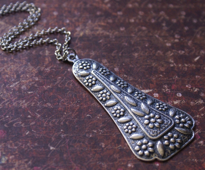 Puede incluir: Un collar con un colgante de plata con un dise&ntilde;o floral detallado. El colgante tiene forma de l&aacute;grima y presenta intrincados tallados de hojas y flores.