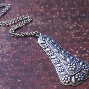 Puede incluir: Un collar de plata con un colgante en forma de &aacute;rbol estilizado con detalles florales. El colgante est&aacute; intrincadamente dise&ntilde;ado con hojas y flores en relieve.