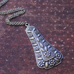 Puede incluir: Un collar de plata con un gran colgante adornado con un dise&ntilde;o de &aacute;rbol estilizado. El colgante est&aacute; detallado con intrincados patrones florales y tiene una superficie ligeramente texturizada.