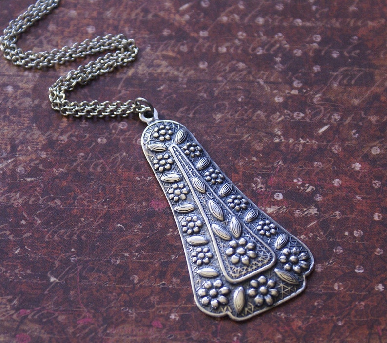 Puede incluir: Un collar de plata con un gran colgante adornado con un dise&ntilde;o de &aacute;rbol estilizado. El colgante est&aacute; detallado con intrincados patrones florales y tiene una superficie ligeramente texturizada.