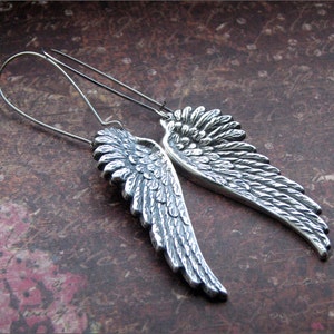 May include: A pair of silver earrings featuring detailed angel wing designs. The wings are textured and have a slightly curved shape.