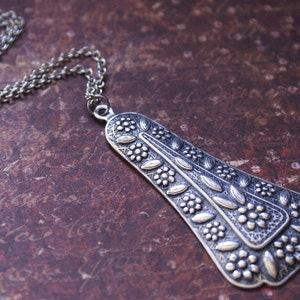 Puede incluir: Un collar de plata con un gran colgante adornado con un dise&ntilde;o floral. El colgante tiene forma de l&aacute;grima y tiene una superficie texturizada.