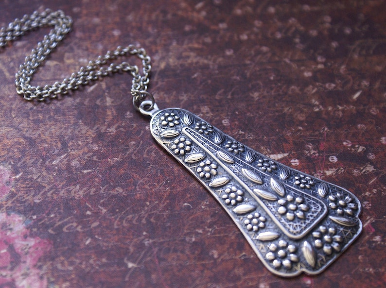 Puede incluir: Un collar de plata con un gran colgante adornado con un dise&ntilde;o floral. El colgante tiene forma de l&aacute;grima y tiene una superficie texturizada.