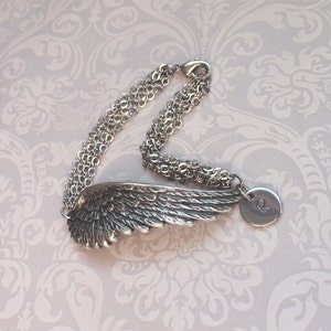 May include: A silver charm bracelet with a detailed angel wing charm and a round charm with the letter "R" engraved on it. The bracelet is made of multiple chains.