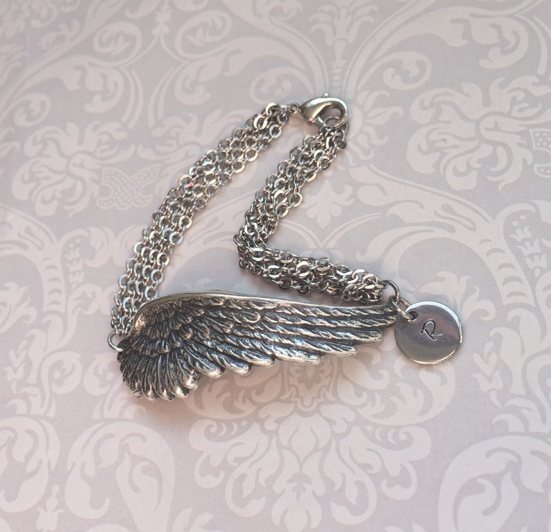 May include: A silver charm bracelet with a detailed angel wing charm and a round charm with the letter "R" engraved on it. The bracelet is made of multiple chains.