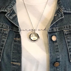 Puede incluir: Un collar de plata con un dije en forma de medallón y un inserto de vidrio transparente. El collar se lleva sobre una camiseta blanca con el texto "courage and" impreso en ella.