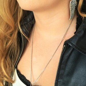 May include: A silver necklace and earrings with a wing design. The necklace is worn by a person with blonde hair and a black leather jacket.