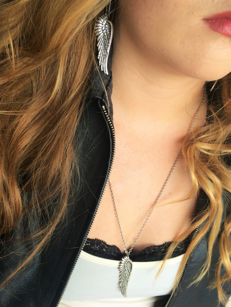 May include: A silver necklace with a wing pendant hangs on a person wearing a black leather jacket and a white shirt. The person is also wearing silver wing earrings.