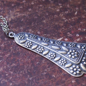 Puede incluir: Un collar de plata con un colgante detallado con un dise&ntilde;o floral. El colgante tiene forma de l&aacute;grima y una superficie texturizada.