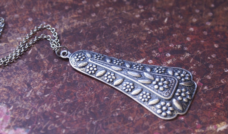 Puede incluir: Un collar de plata con un colgante detallado con un dise&ntilde;o floral. El colgante tiene forma de l&aacute;grima y una superficie texturizada.