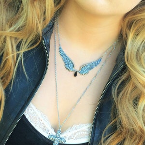 May include: A woman wearing a black leather jacket with a silver chain necklace featuring a cross pendant and a pair of angel wings with a black gemstone.