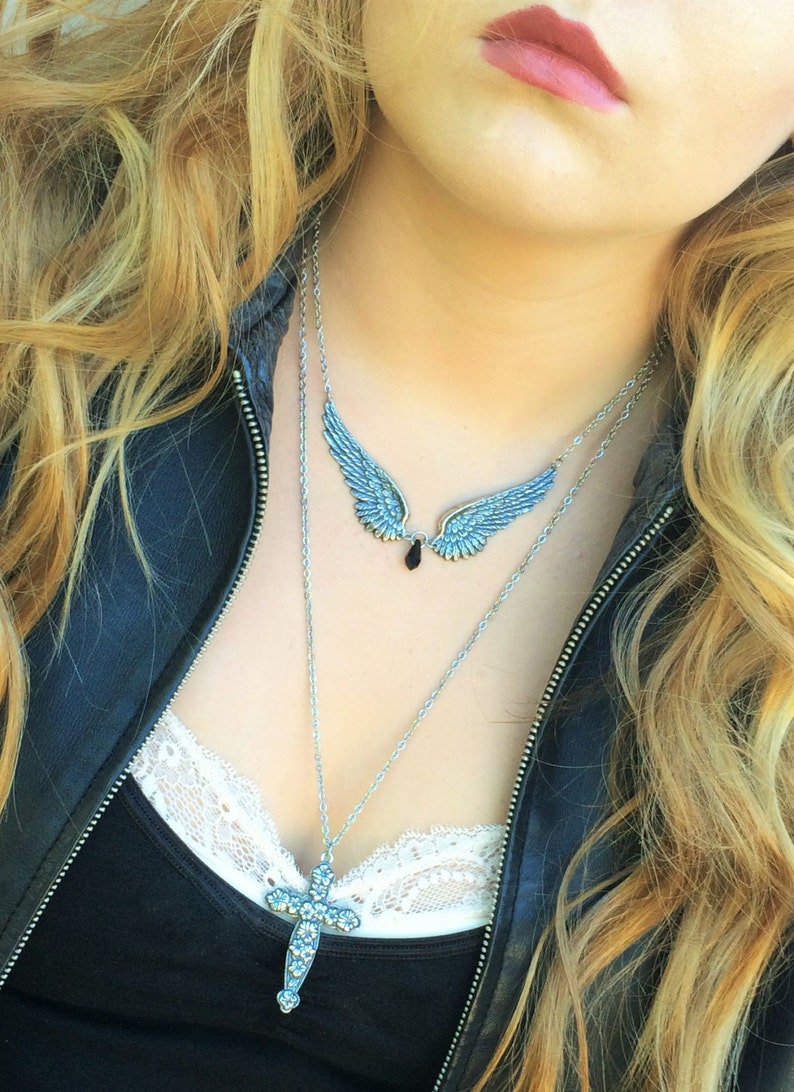 May include: A woman wearing a black leather jacket with a silver chain necklace featuring a cross pendant and a pair of angel wings with a black gemstone.
