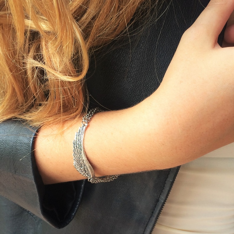 Puede incluir: Un brazalete de plata en forma de ala con un detalle de cadena. El brazalete se lleva en la mu&ntilde;eca.