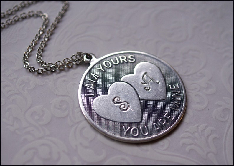 May include: A silver-toned pendant necklace with a chain. The pendant is round with the words "I AM YOURS YOU ARE MINE" around the edge. Two heart shapes are in the center, each with a letter. One heart has an "S" and the other has an "A".