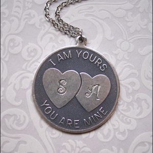 May include: A silver-toned pendant necklace with a chain. The round pendant features the engraved text "I AM YOURS YOU ARE MINE". Two hearts are in the center, each with a letter. The letter "S" is in the left heart and the letter "A" is in the right heart.