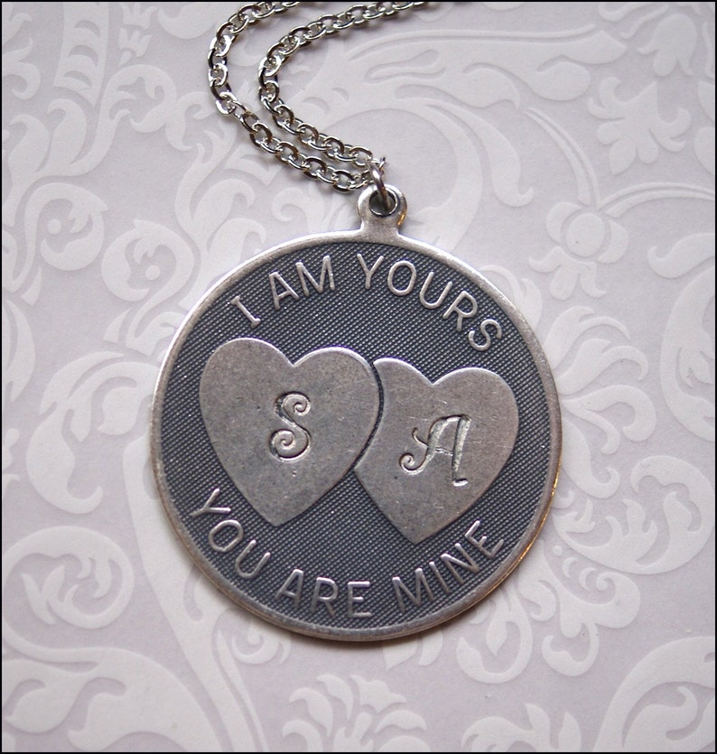 May include: A silver-toned pendant necklace with a chain. The round pendant features the engraved text "I AM YOURS YOU ARE MINE". Two hearts are in the center, each with a letter. The letter "S" is in the left heart and the letter "A" is in the right heart.