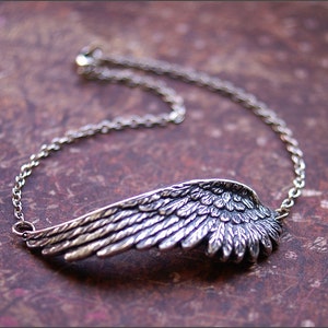 Könnte beinhalten: Ein silbernes Kettenarmband mit einem detaillierten Flügelanhänger. Der Flügel ist aus Metall gefertigt und hat eine strukturierte Oberfläche.