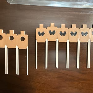 May include: A collection of tan paper targets with white sticks. Some targets have circular holes, while others feature a black "W" or a handprint design. The targets are arranged on a dark brown surface.