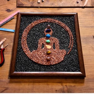 May include: A square framed wall art piece featuring a mosaic of a meditating figure in copper-colored stones against a black stone background. The figure is adorned with seven small, smooth, multi-colored stones aligned vertically along the center.