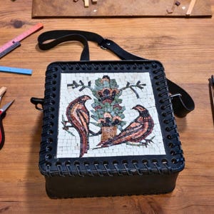 May include: A square black leather shoulder bag featuring a central mosaic panel depicting two birds perched on branches flanking a stylized tree. The bag has a stitched border and a black strap, surrounded by various metal crafting tools.