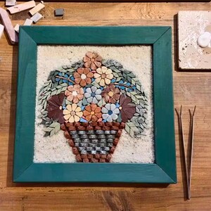 May include: A mosaic artwork of a flower basket in a green frame. The mosaic features various colors of tiles, including brown, orange, blue, and green. Crafting tools, including pliers and tweezers, are arranged around the artwork on a wooden surface.