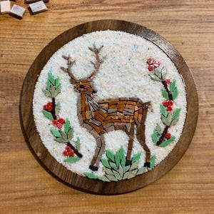 May include: A round, mosaic art piece featuring a deer with antlers, crafted from brown and tan tiles. The deer is surrounded by green leaves and red berries, set against a white background, within a wooden frame.