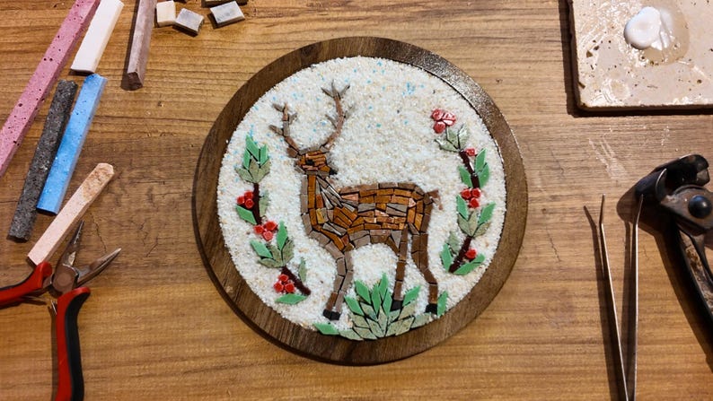 May include: A round, mosaic art piece featuring a deer with antlers, crafted from brown and tan tiles. The deer is surrounded by green leaves and red berries, set against a white background, within a wooden frame.