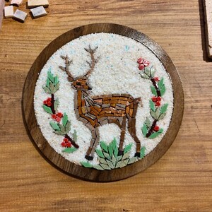 May include: A mosaic art piece featuring a deer with antlers, crafted from brown and tan tiles, surrounded by green leaves and red berries. The artwork is set in a round wooden frame. Various mosaic tools and materials are scattered around the piece.