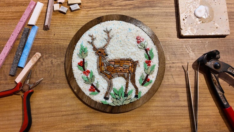 May include: A mosaic art piece featuring a deer with antlers, crafted from brown and tan tiles, surrounded by green leaves and red berries. The artwork is set in a round wooden frame. Various mosaic tools and materials are scattered around the piece.