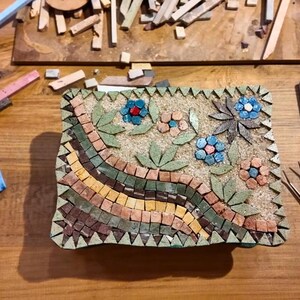 May include: A rectangular mosaic box featuring a floral pattern with blue and copper-colored petals, green leaves, and a wavy border of brown, yellow, and green tiles. The box is surrounded by metal pliers, tweezers, and various loose stone tiles.