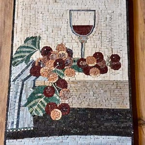 May include: Mosaic artwork depicting a wine glass filled with red wine, grapes, and leaves. The mosaic is made of small, square tiles in shades of white, brown, and red. The artwork is framed in a black border.