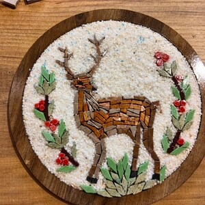 May include: A mosaic art piece featuring a deer with antlers, crafted from various brown and tan tiles. The deer is set against a white background within a round wooden frame. Green leaves and red berries surround the deer.
