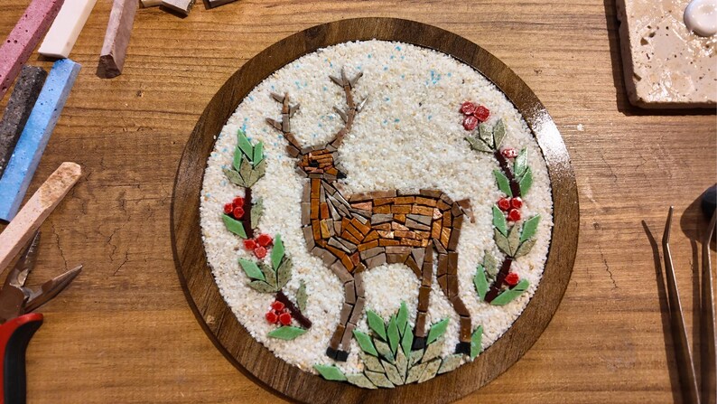 May include: A mosaic art piece featuring a deer with antlers, crafted from various brown and tan tiles. The deer is set against a white background within a round wooden frame. Green leaves and red berries surround the deer.