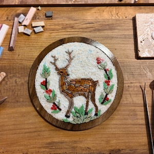 May include: A handcrafted mosaic art piece featuring a deer design, surrounded by floral elements. The mosaic is set within a round wooden frame. Various tools and mosaic tiles in shades of pink, blue, and beige are scattered around the artwork.