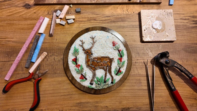 May include: A handcrafted mosaic art piece featuring a deer design, surrounded by floral elements. The mosaic is set within a round wooden frame. Various tools and mosaic tiles in shades of pink, blue, and beige are scattered around the artwork.
