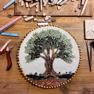 May include: A circular mosaic art piece depicting a tree with green leaves and a brown trunk, set against a white background. The artwork is surrounded by various crafting tools, including red-handled pliers, metal tweezers, and scattered stone tiles.