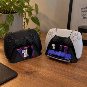 Puede incluir: Dos mandos de videojuegos, uno negro y otro blanco, se muestran sobre soportes negros. Cada soporte presenta el logotipo de Grand Theft Auto VI. El soporte del mando blanco incluye un pequeño modelo de coche de juguete azul en la base.