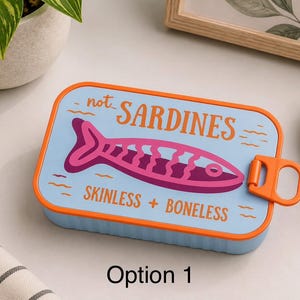 Può includere: Un contenitore rettangolare azzurro con bordo arancione e coperchio a linguetta. Il coperchio presenta una grafica stilizzata di un pesce viola con il testo "not SARDINES" e "SKINLESS + BONELESS" stampato in arancione, con piccole icone a onda.