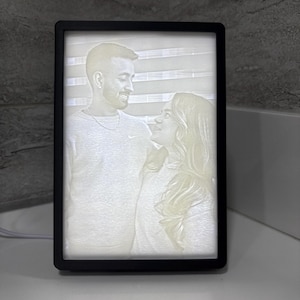 Peut inclure: Un cadre lumineux à LED noir affichant une image en rendu 3D d'un couple. L'image est en niveaux de gris, montrant le couple se regardant. Le cadre lumineux est rectangulaire et possède un cordon d'alimentation blanc.