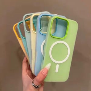 May include: A selection of pastel-coloured phone cases in yellow, teal, light blue, and green. Each case has a contrasting border around the camera and a white circular design on the back. The cases are held in a hand.