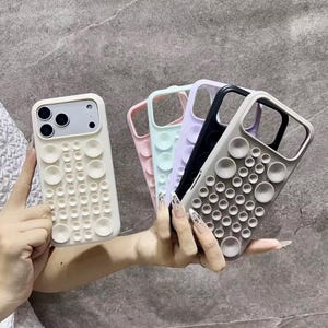 Puede incluir: Una colección de fundas de teléfono coloridas con un diseño de burbujas único. Las fundas están disponibles en varios colores, incluyendo blanco roto, rosa, azul claro, morado, negro y gris. Las fundas tienen una superficie texturizada con elementos circulares en relieve.